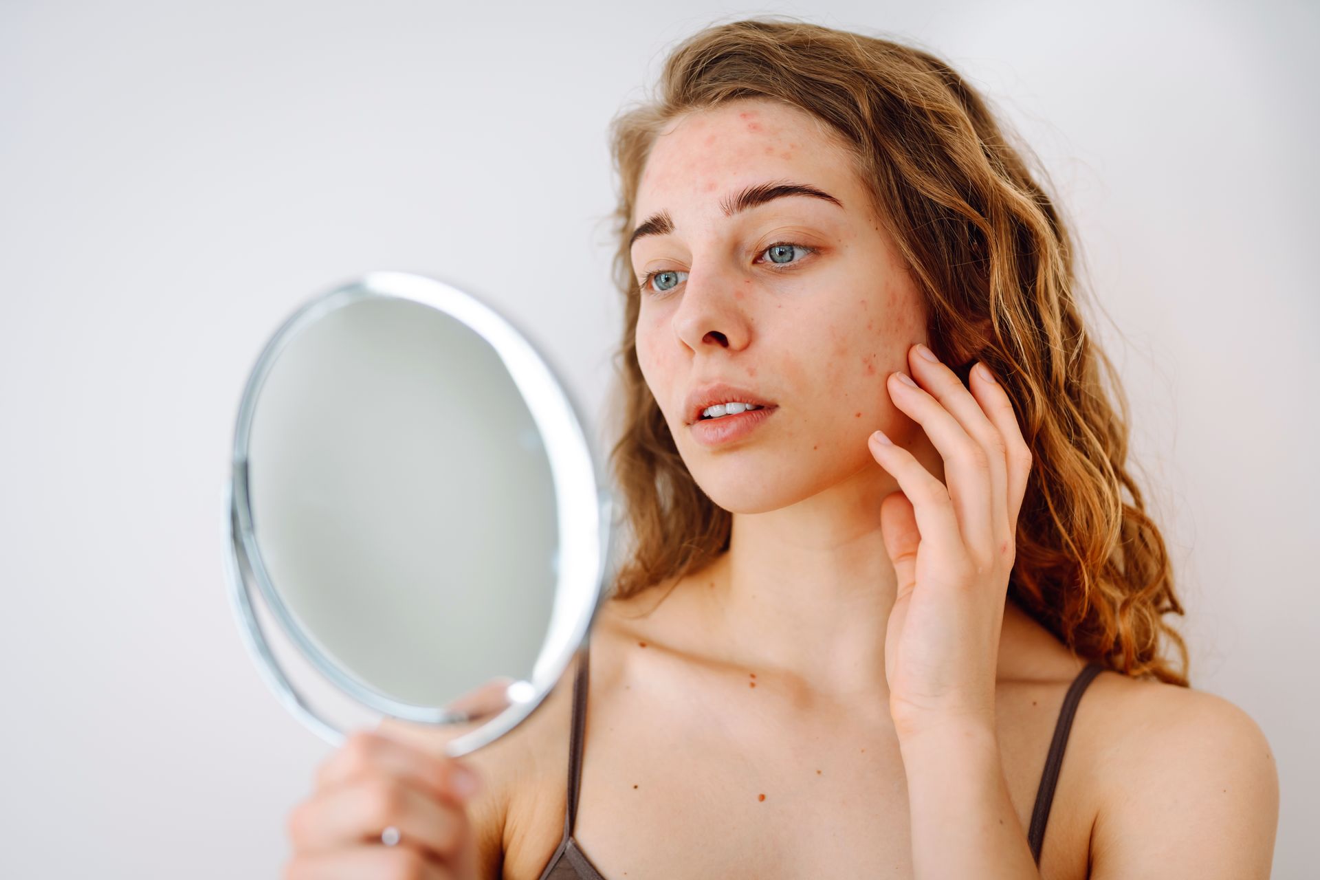Woman looking at her face in a mirror, touching her cheek. Indoor setting with a white background. She appears concerned.