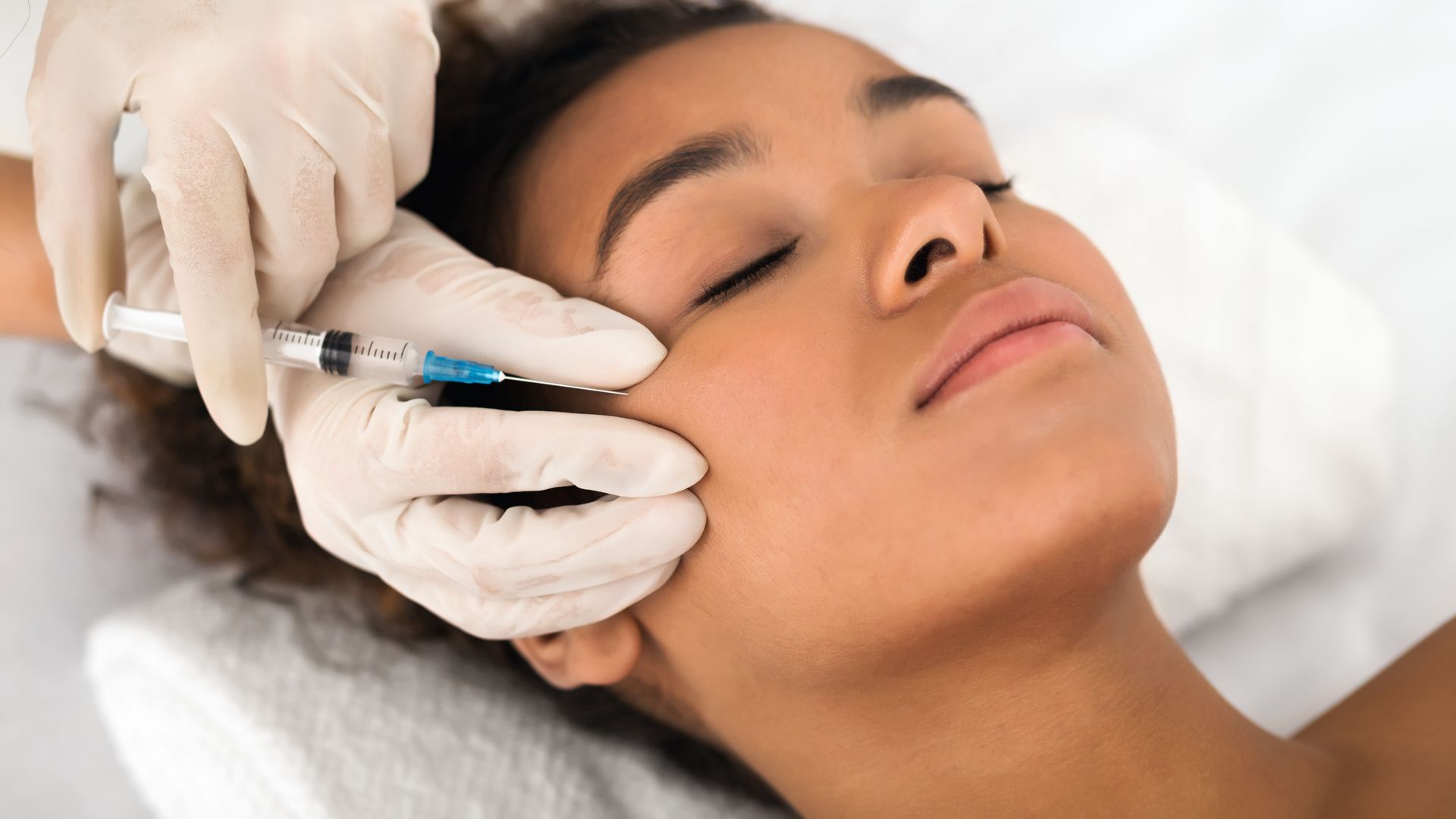A person receiving an injection in their cheek; gloved hand holding syringe near face.