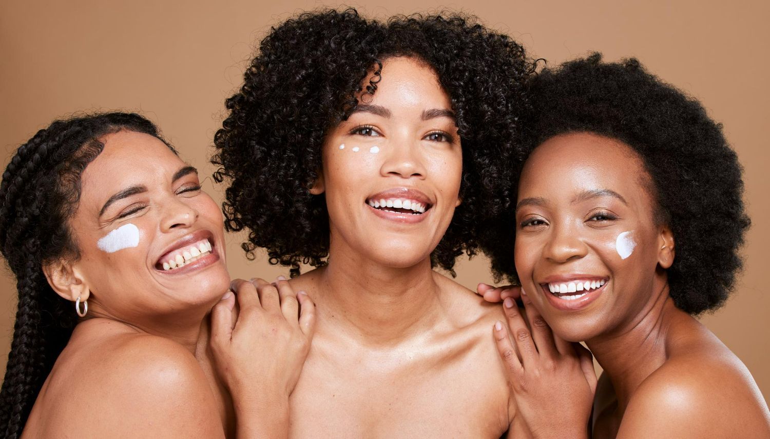 Three smiling people posing closely together against a warm beige background, one with white cream on their face and shoulder