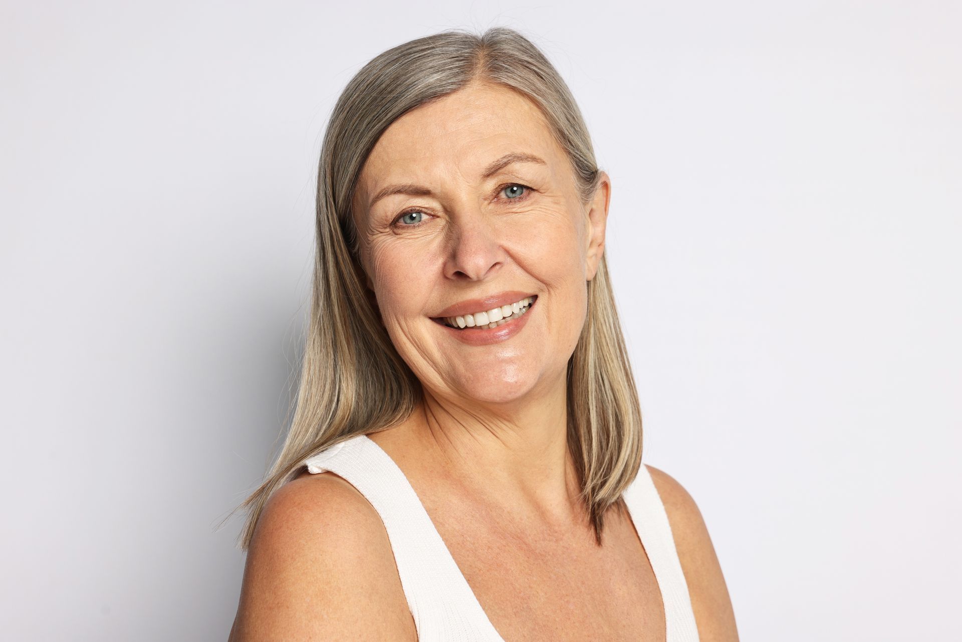 Smiling woman with shoulder-length gray hair in a white sleeveless top against a light gray background