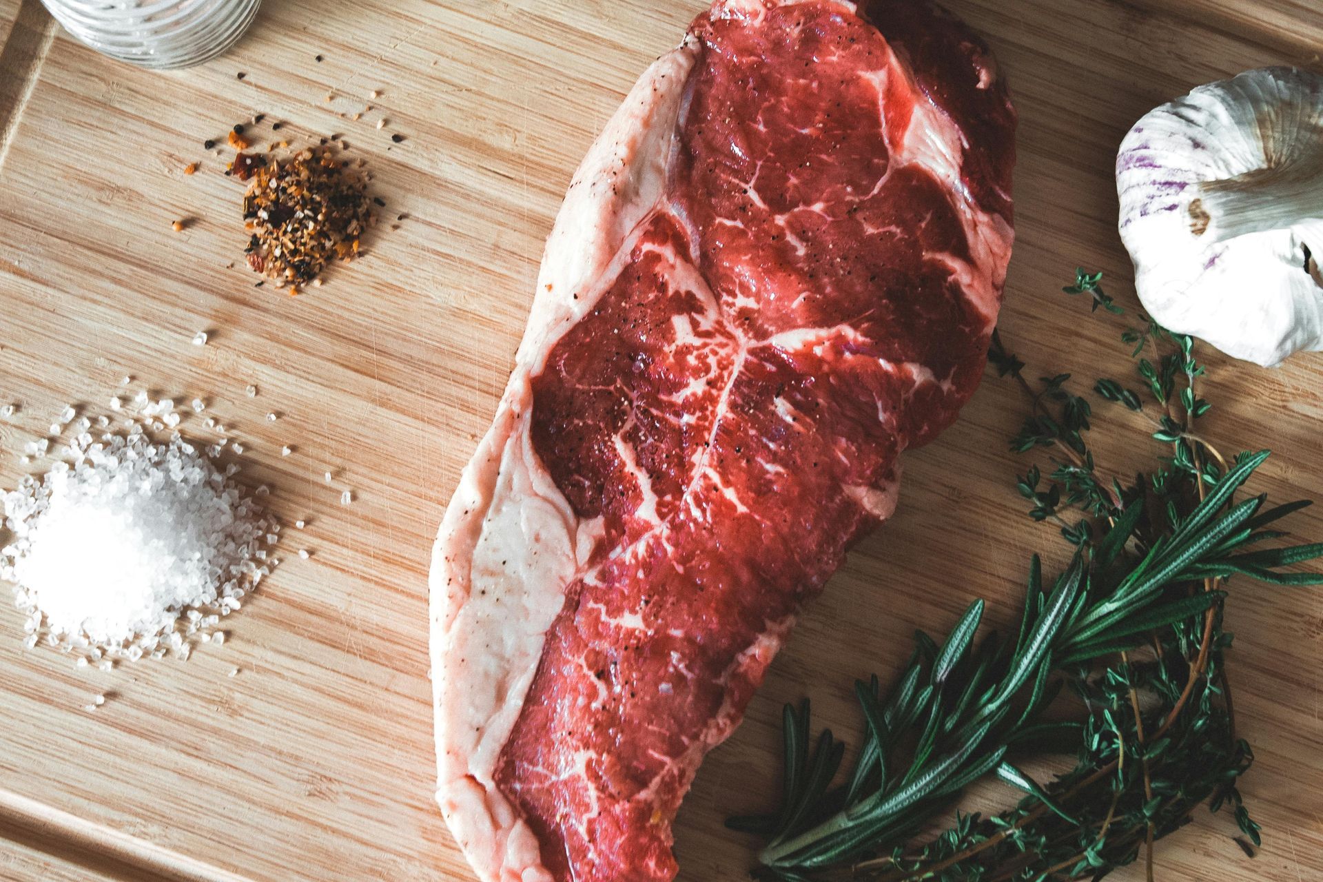 Raw ribeye steak on a wooden cutting board with salt, spices, and rosemary.