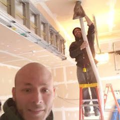 A man is standing on a ladder in a garage while another man is working on the ceiling.