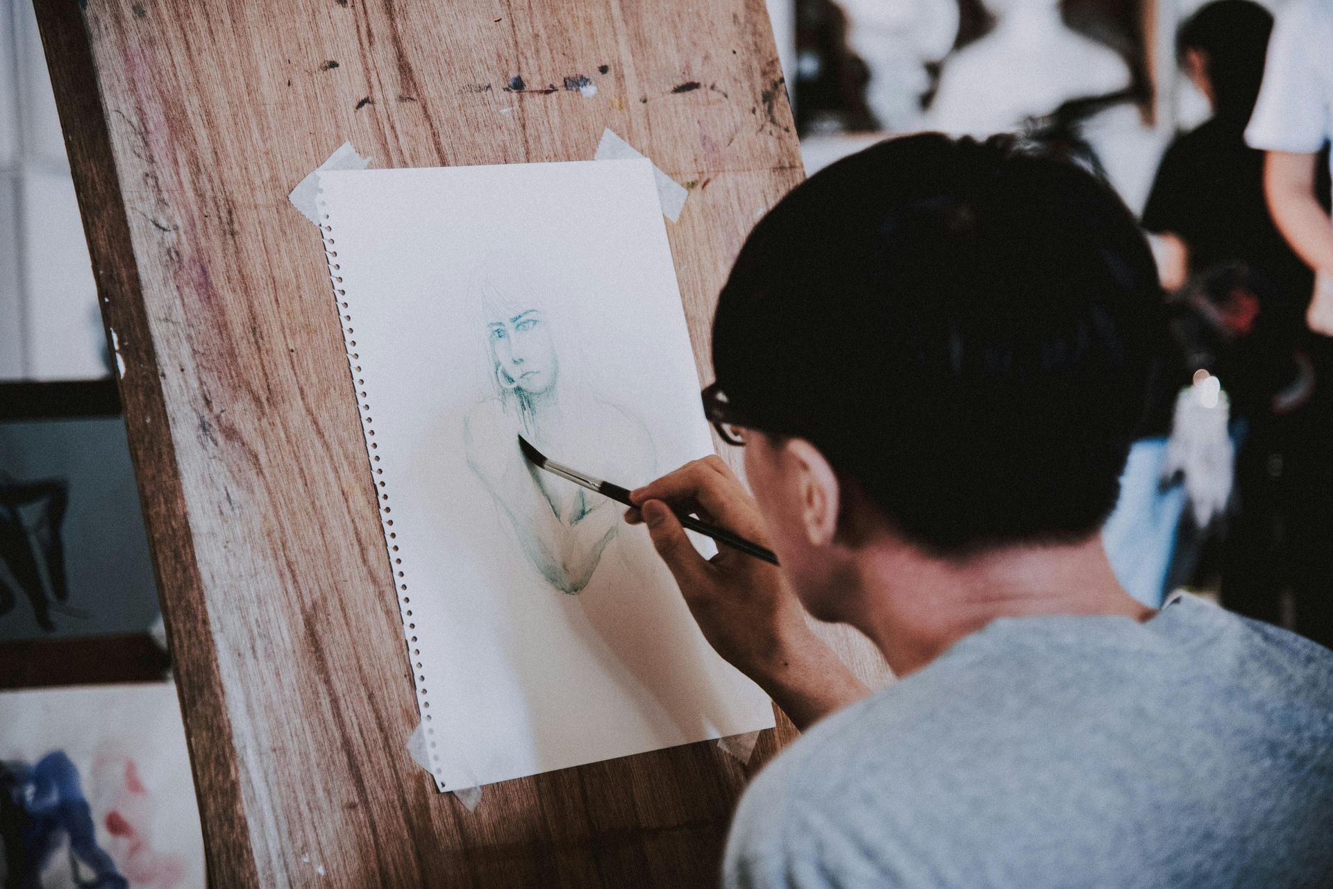 Artist sketching a figure with a paintbrush, paper taped to a wooden easel. Other artwork and people in the background.