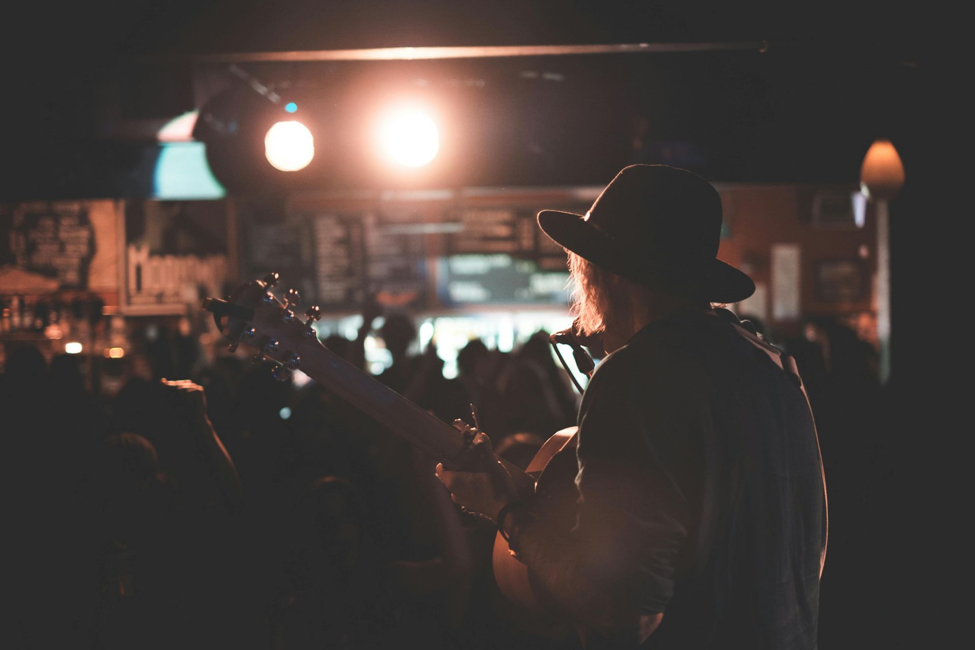 Guitarist in a hat performing on stage at a dimly lit bar, silhouetted against a bright spotlight.