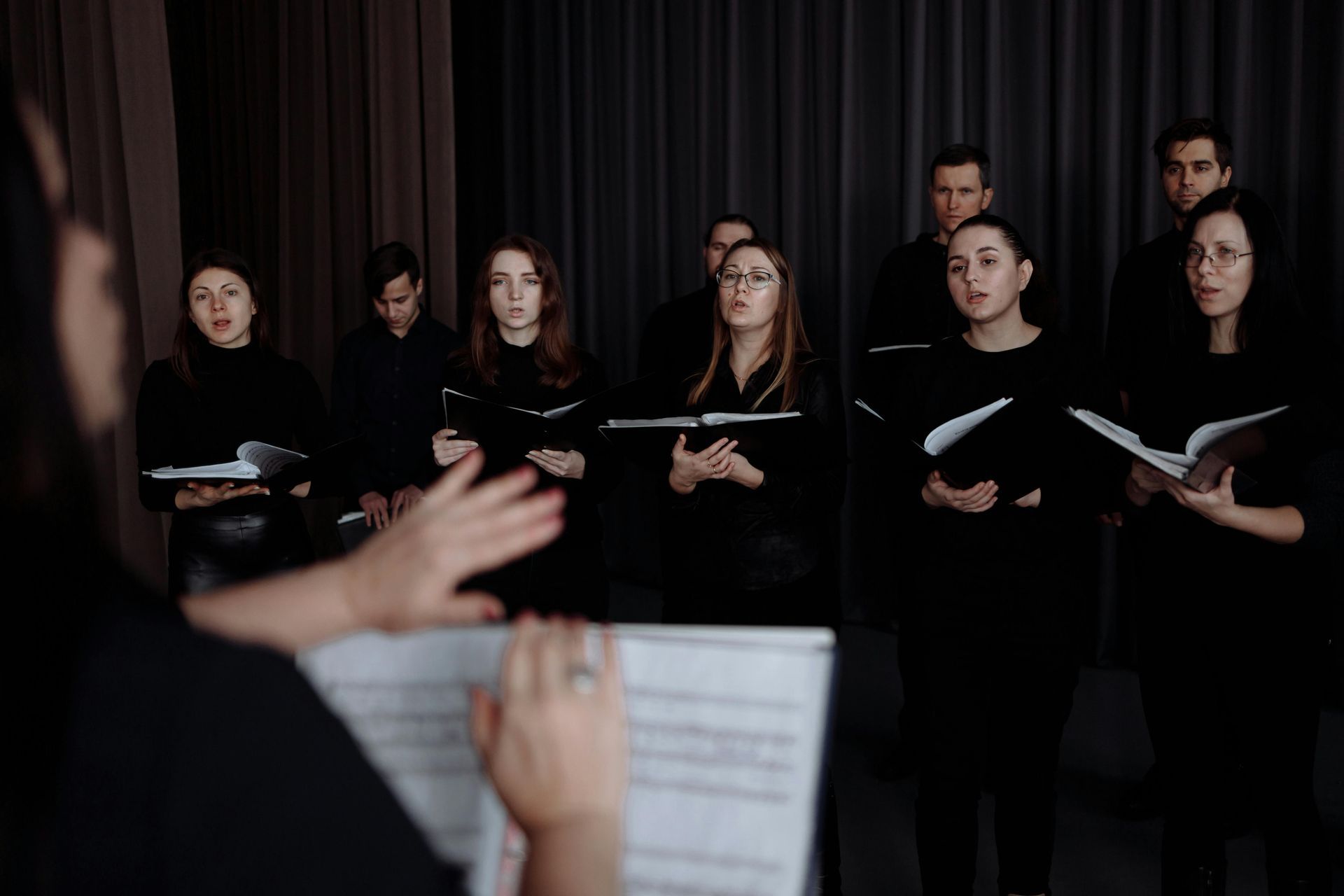 Choir rehearsing, conductor with sheet music, singers in dark clothing hold music books, gray background.