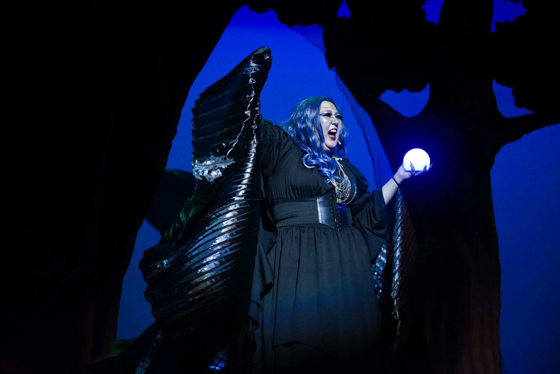 Woman in dark costume, holding glowing orb. Forest backdrop, blue hair, singing with open mouth.