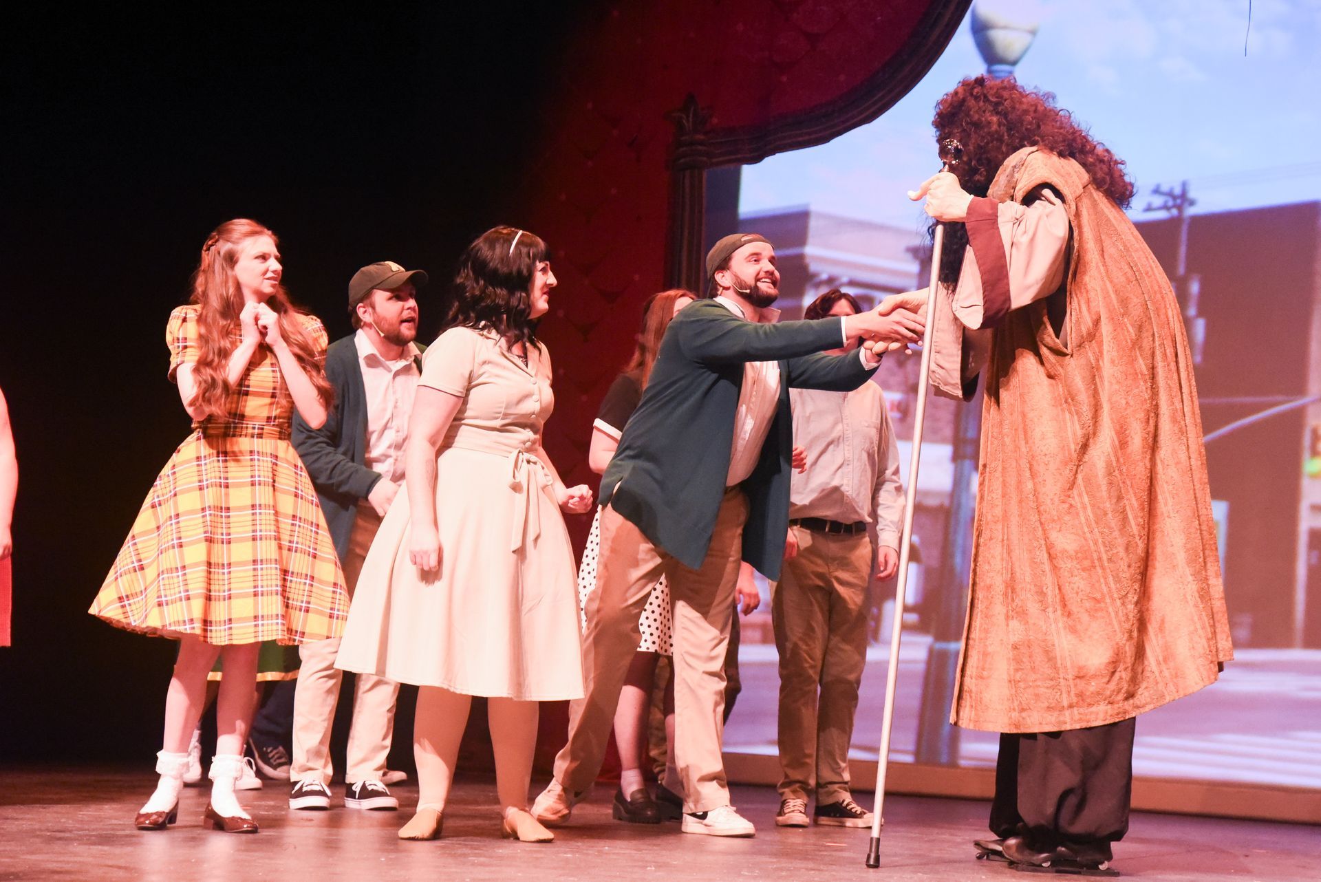 A stage performance. Cast greets a tall figure in a robe. Audience looks on.