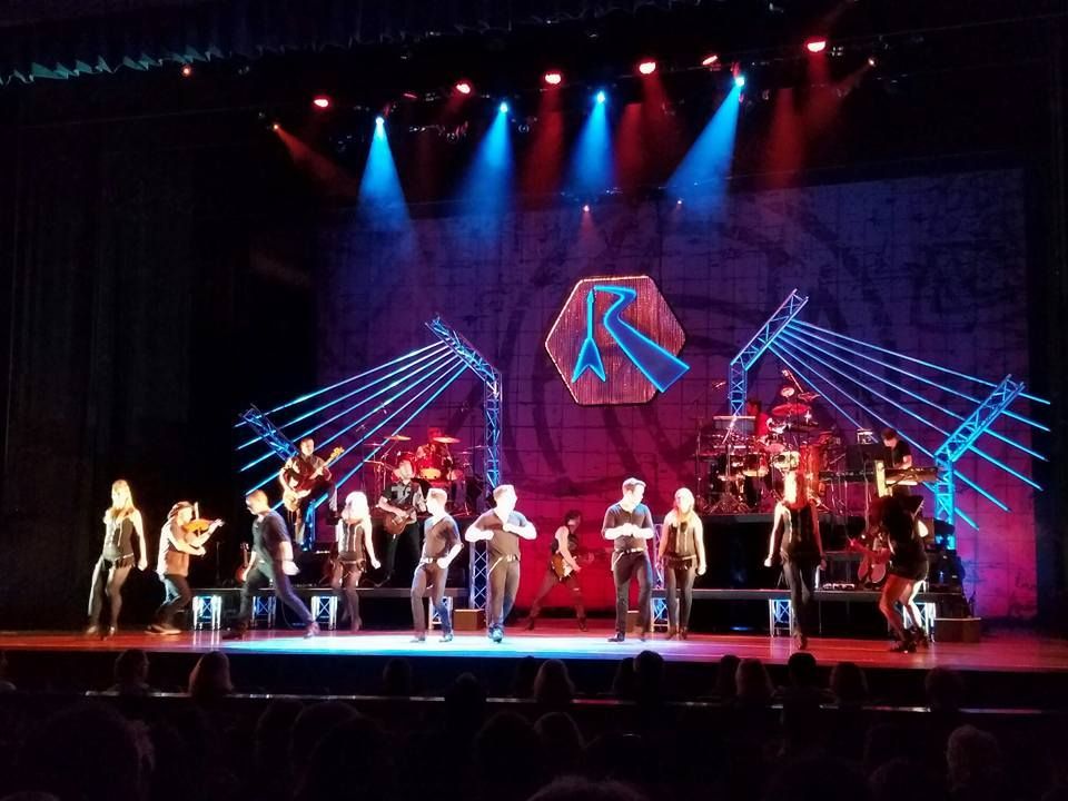 A stage performance with performers in black, a band, and a logo with blue and red lighting.