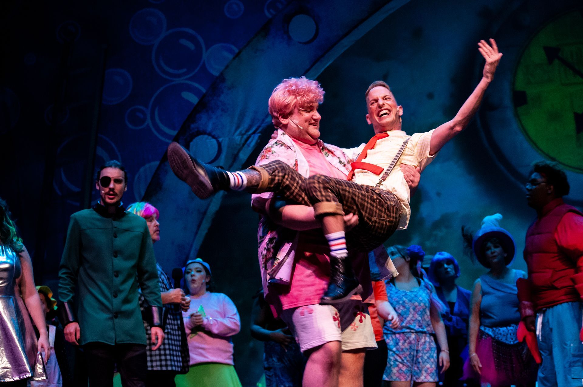 A theatrical stage scene with a man carrying another, surrounded by actors in colorful costumes.