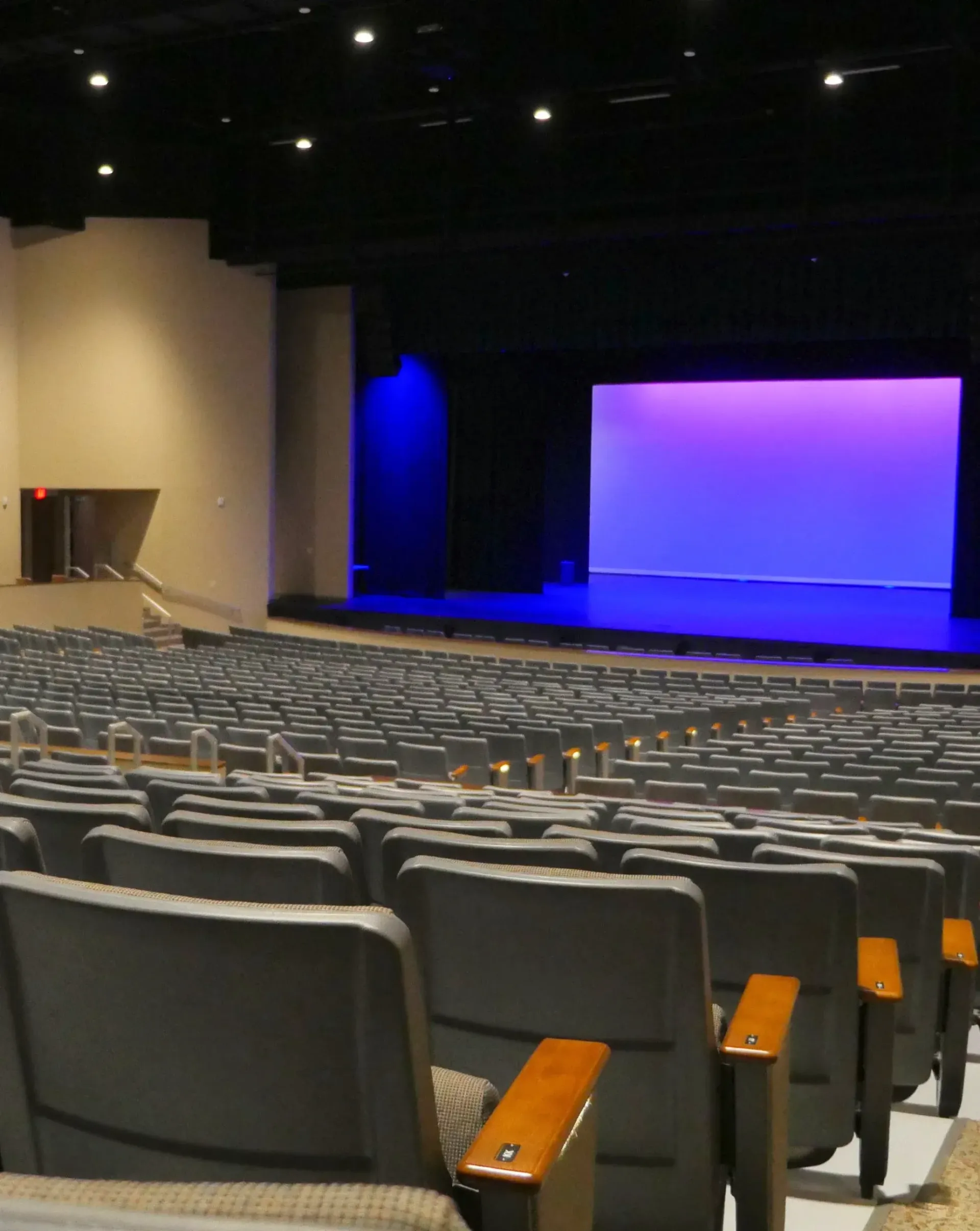 Auditorium interior with rows of gray seats facing a stage with a blue and purple backdrop.