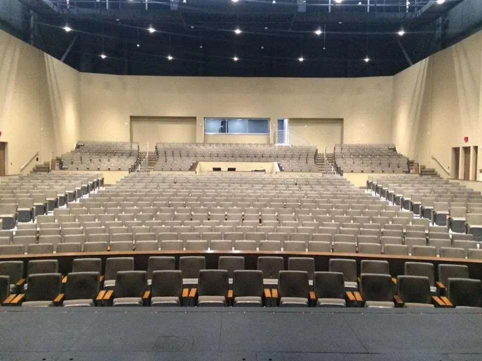 Empty auditorium with rows of seats facing a stage. Beige walls, black ceiling, and dim lighting.
