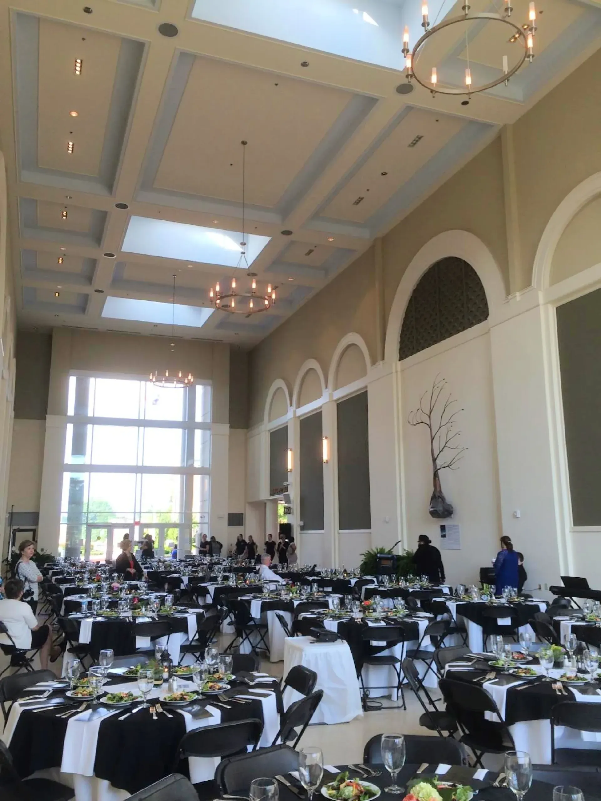 Large banquet hall with black and white tables, high ceilings, chandeliers, and large windows.