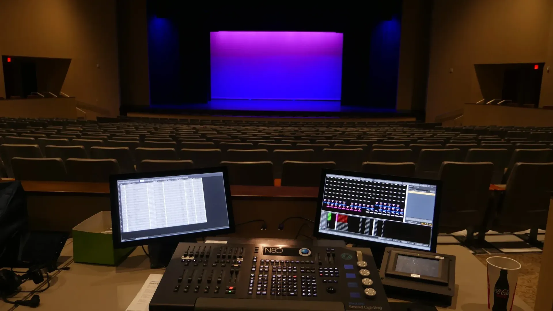 Control board and monitors in a theater, stage with blue and purple lighting, empty seats.