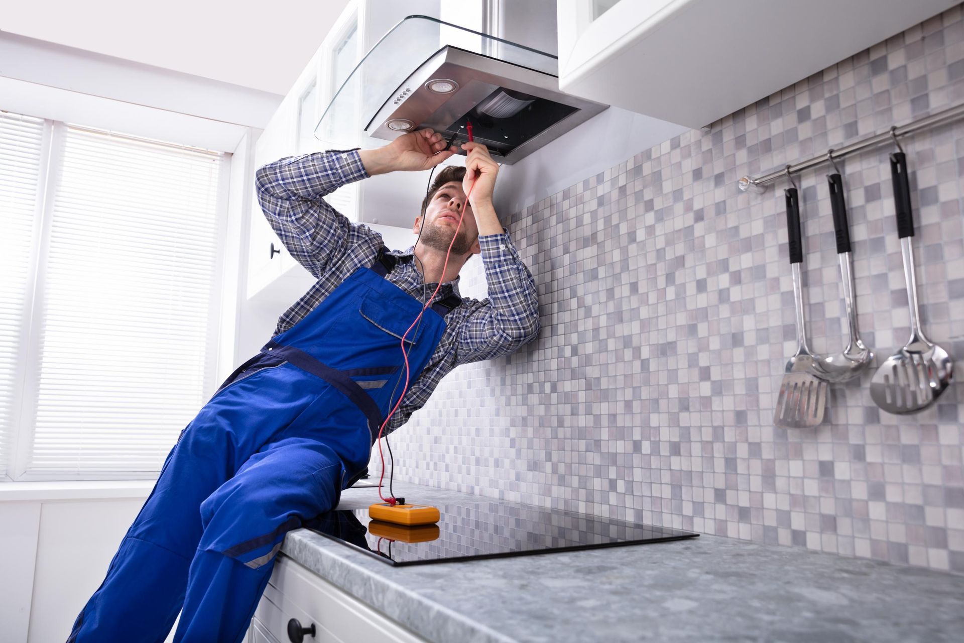 Man in blue overalls working on a kitchen range hood above a countertop, near a window.