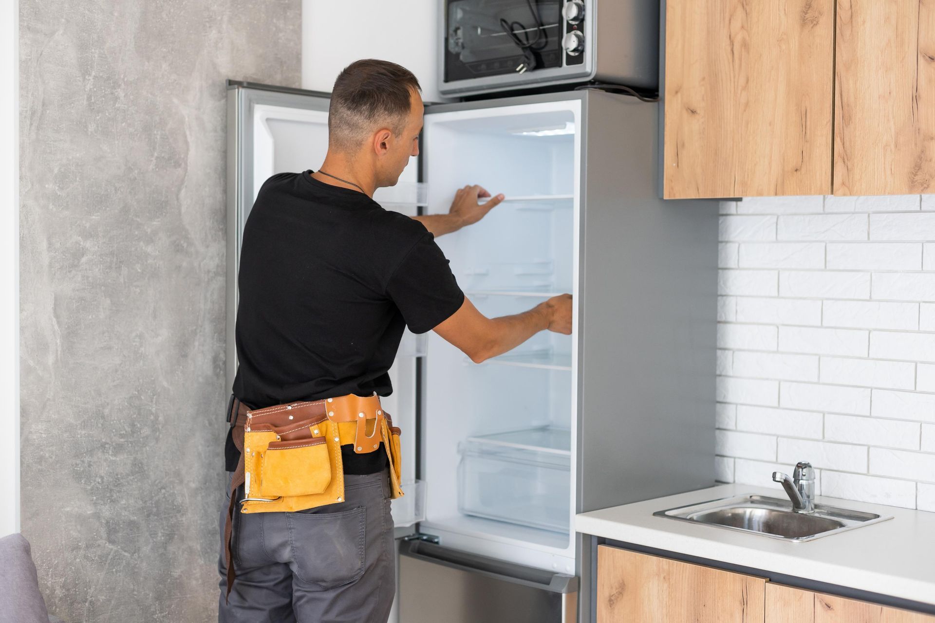 Man with tool belt inspecting the inside of a refrigerator in a kitchen.