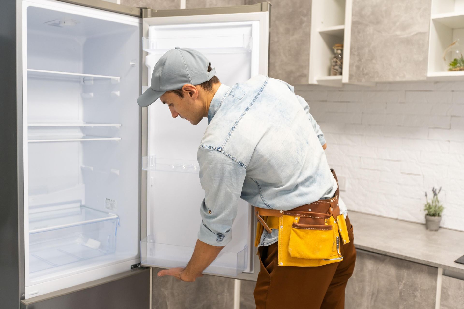 Man in work clothes examining an empty refrigerator in a kitchen.