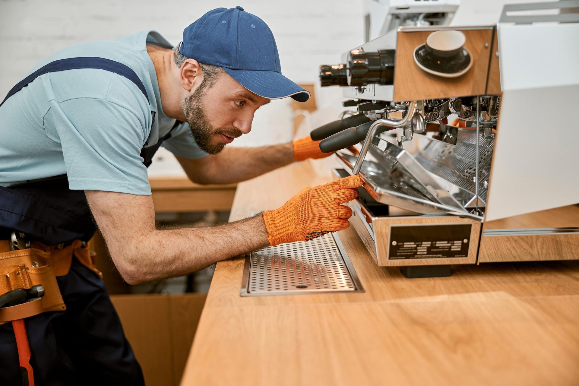 Man in blue cap and gloves repairs espresso machine in a cafe.
