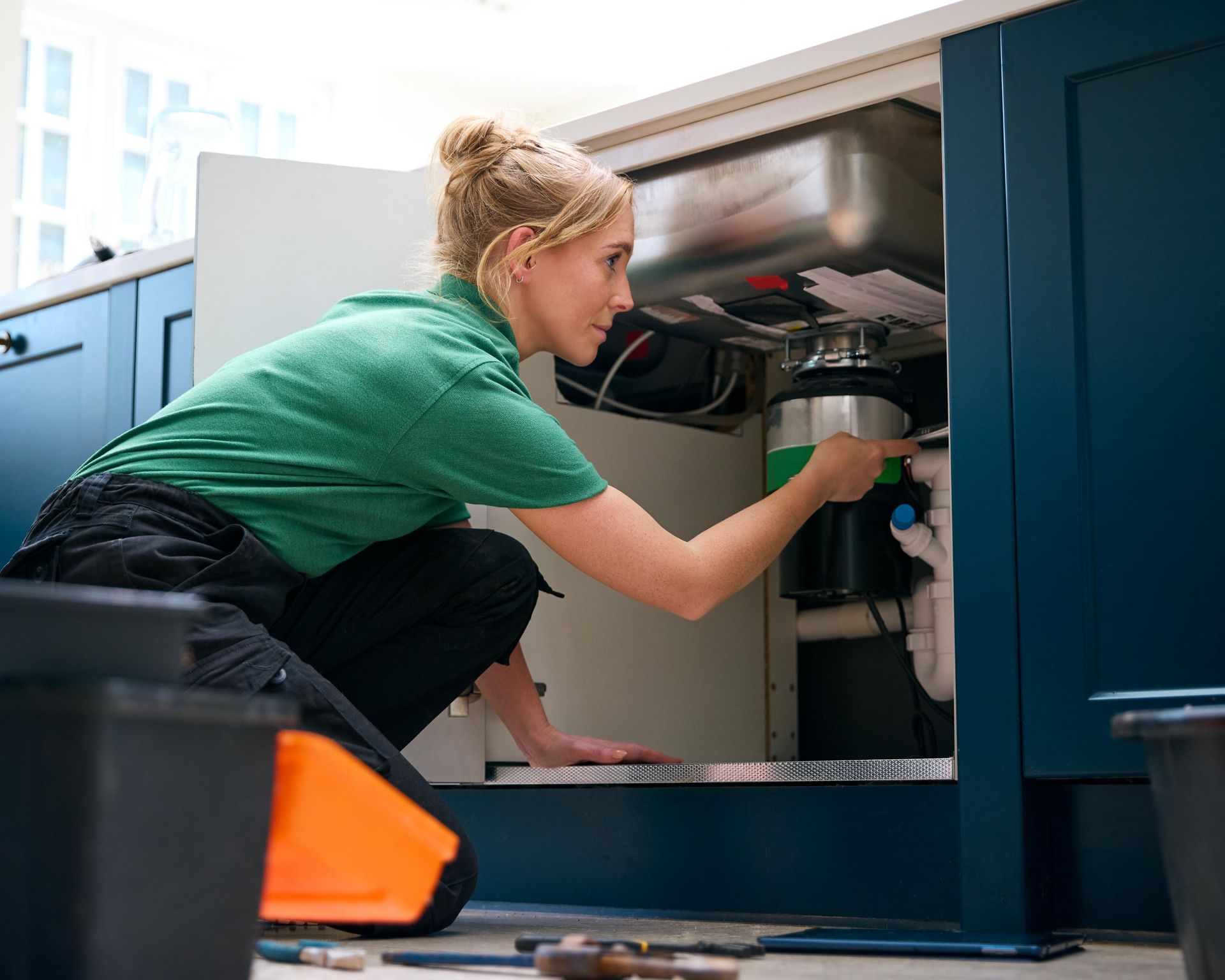 Woman plumber fixing a garbage disposal under a kitchen sink. She is kneeling, wearing green shirt, and using a wrench.