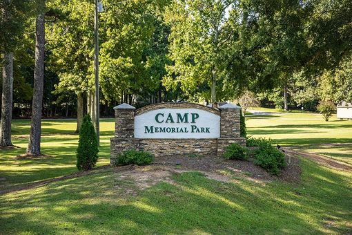 A sign for camp memorial park is surrounded by trees and grass.