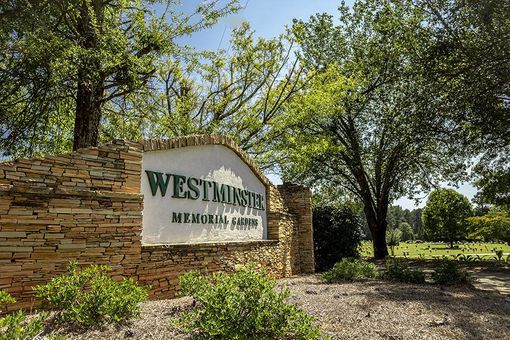 A sign for westminster memorial park is surrounded by trees and bushes.