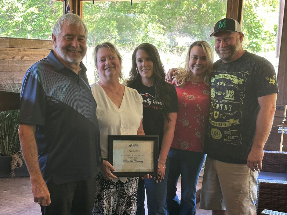 Five people posing outdoors, holding a framed award certificate, with trees and a wooden shelter behind them.