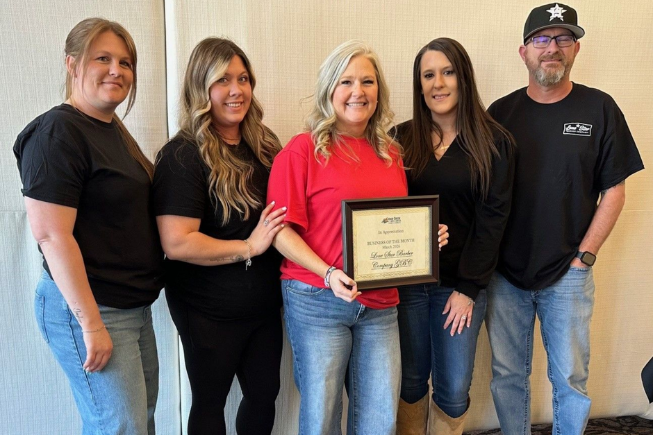 Five people stand in a line against a neutral backdrop. A person in a red shirt holds an award certificate.
