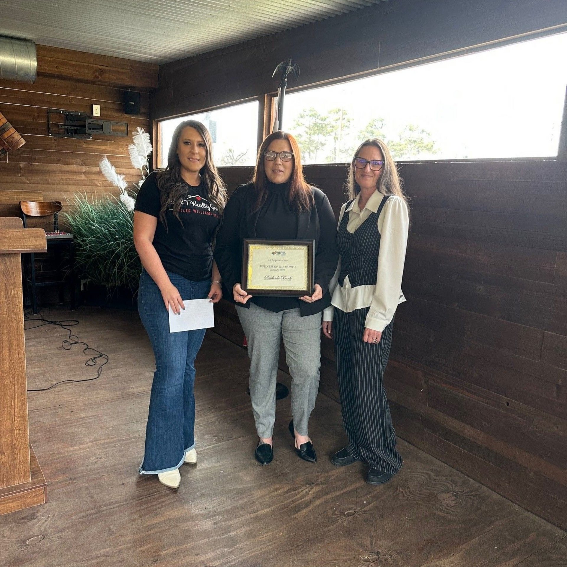 Three women in a room, one holding a certificate. Wooden interior, large windows, and a podium.