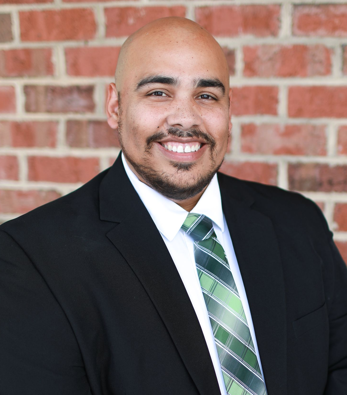 A bald man with a beard wearing a suit and tie is smiling in front of a brick wall.
