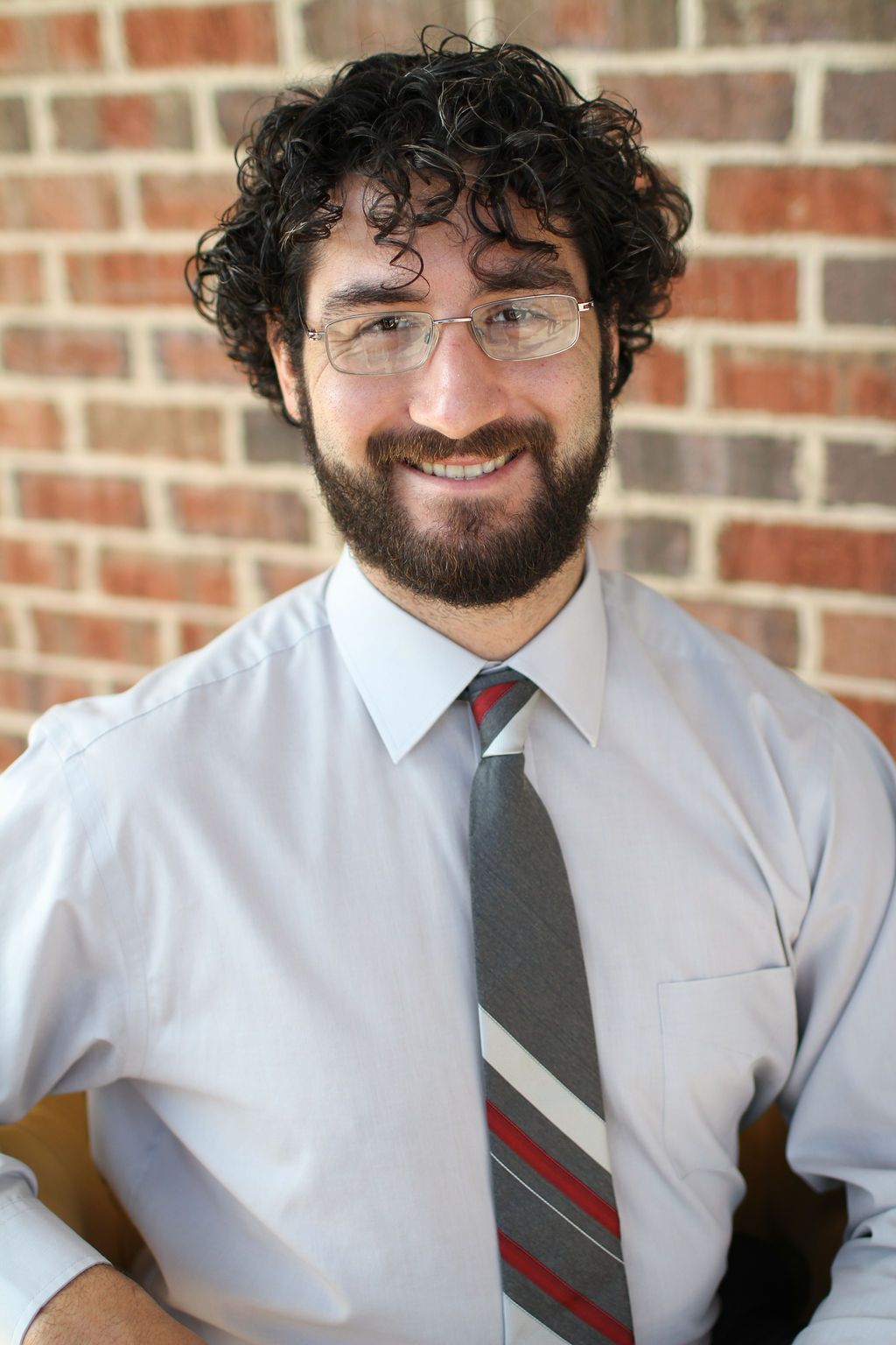 A man with a beard wearing glasses and a tie is smiling in front of a brick wall.