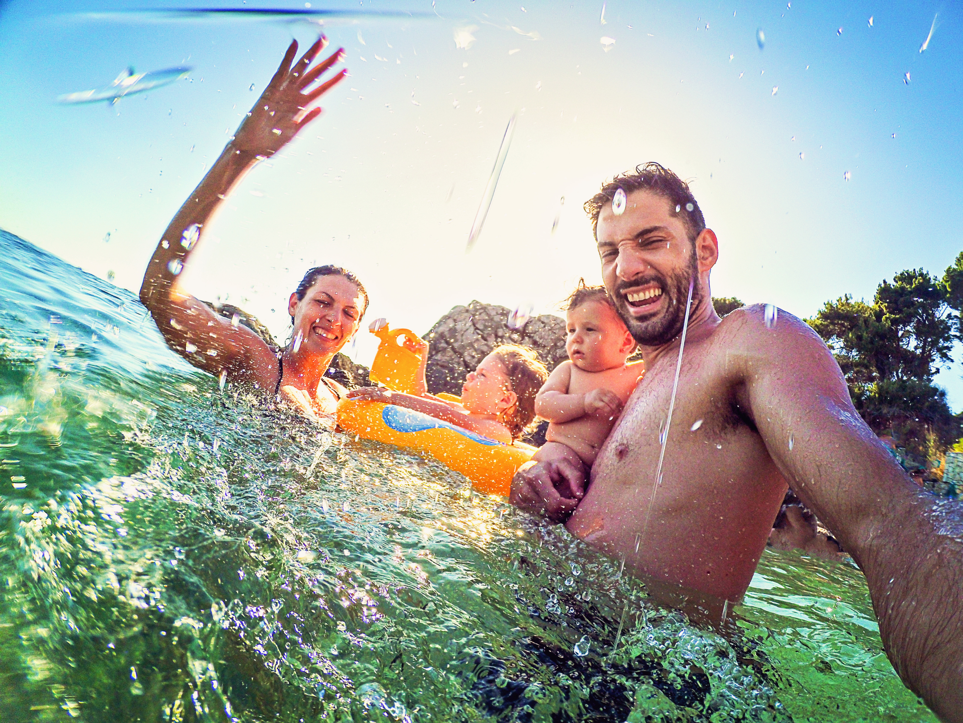 Family in ocean, enjoying a sunny day. Woman waves, man holds baby, other child on float.