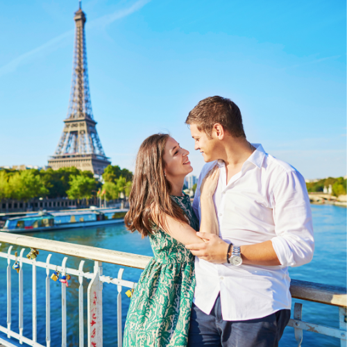 Newlyweds walk on a beach at sunset; bride in white dress, groom in tuxedo; orange sky reflects in turquoise water.