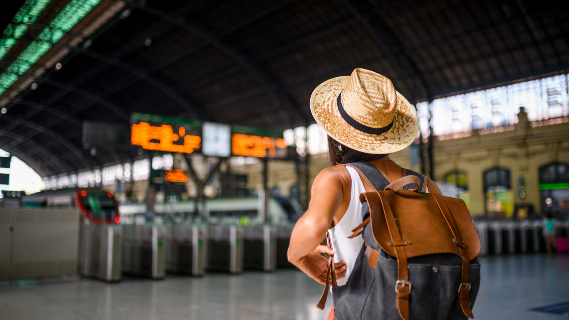 Person with straw hat and backpack at a train station, looking at departure boards.