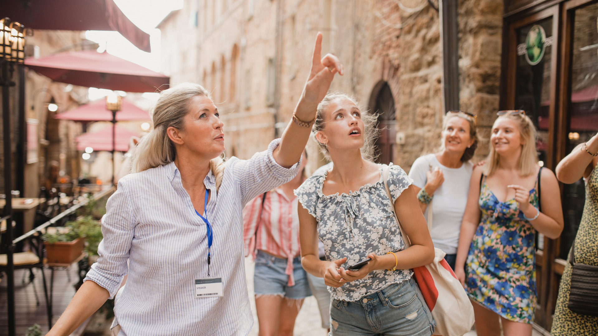 Tour guide pointing upward, leading a group of women on a city street; buildings, shops, and umbrellas in background.