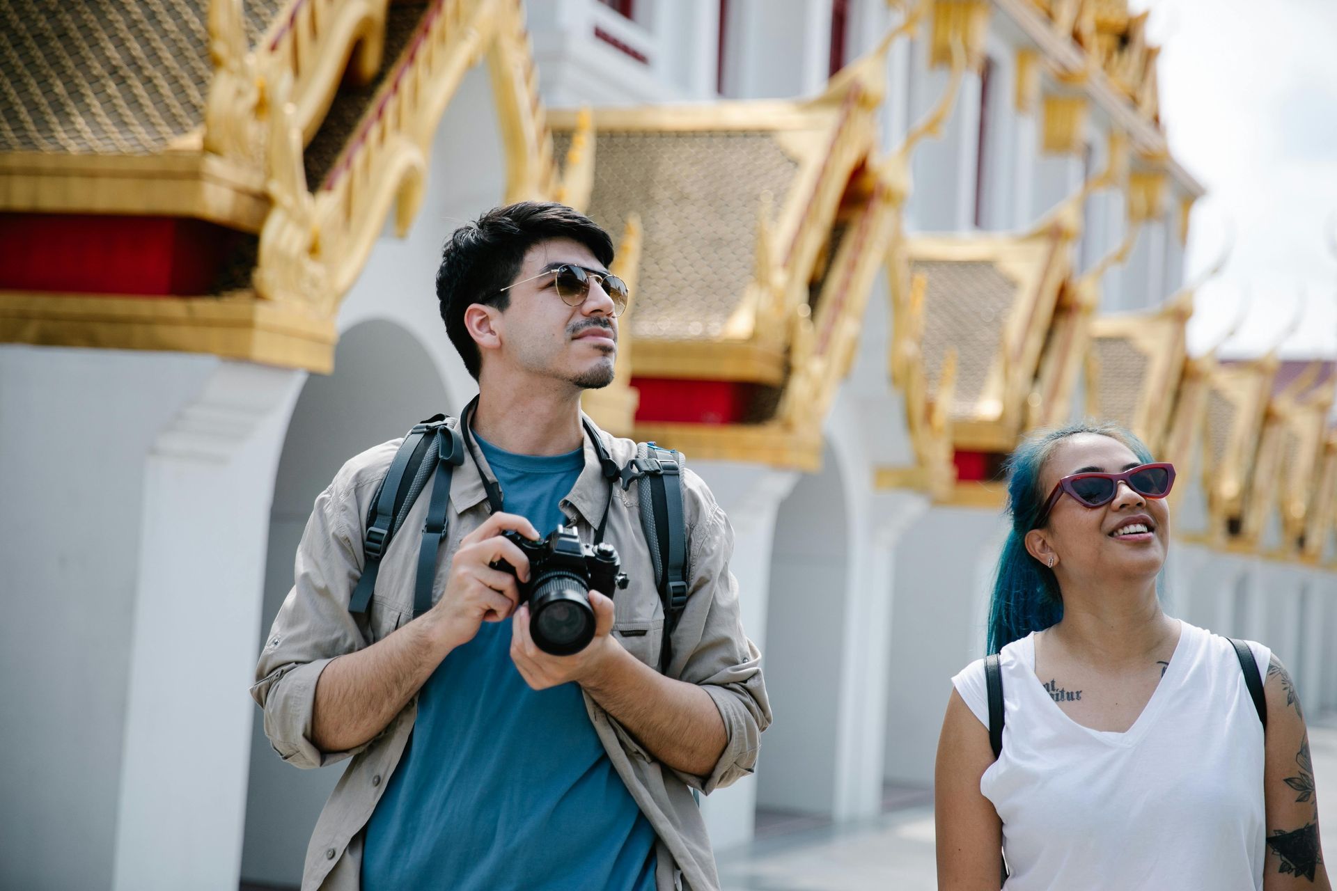 Two tourists with sunglasses, looking up at a gold and white ornate building; man holding camera.