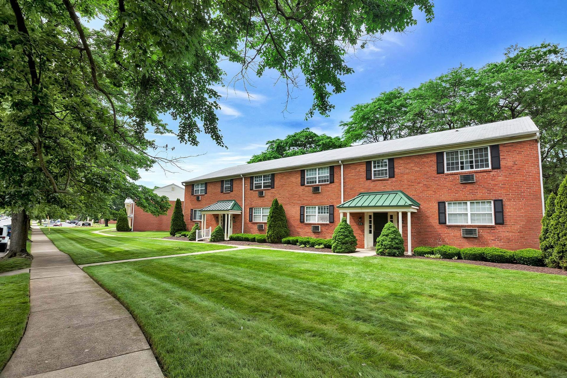 Exterior view of a brick apartment building with a green lawn, trees, and small entry porches.