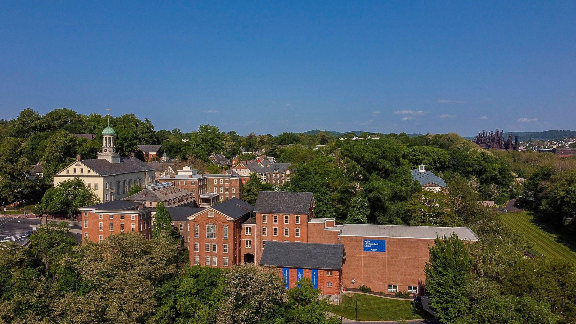 Aerial view of a brick building complex with lush trees and a clear blue sky.