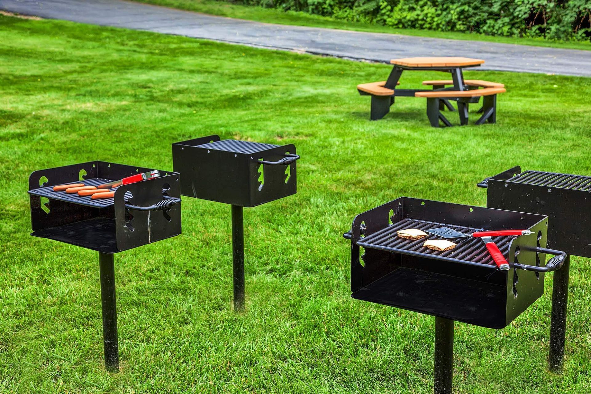 Outdoor community grills on a grassy lawn with a picnic table in the background.