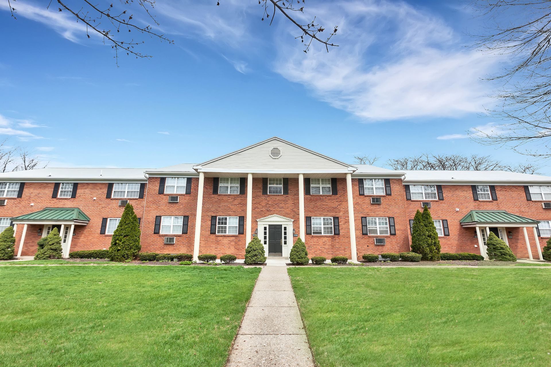 Brick apartment building exterior with central entry, white columns, and manicured lawn.