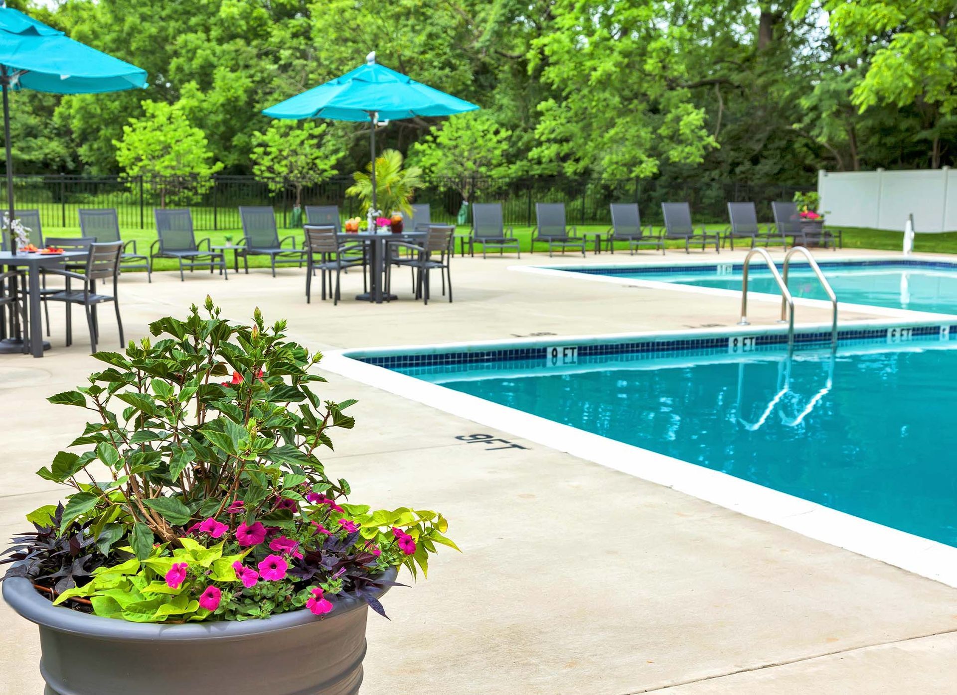 Outdoor pool area with blue umbrellas, lounge chairs, and tables among green trees.
