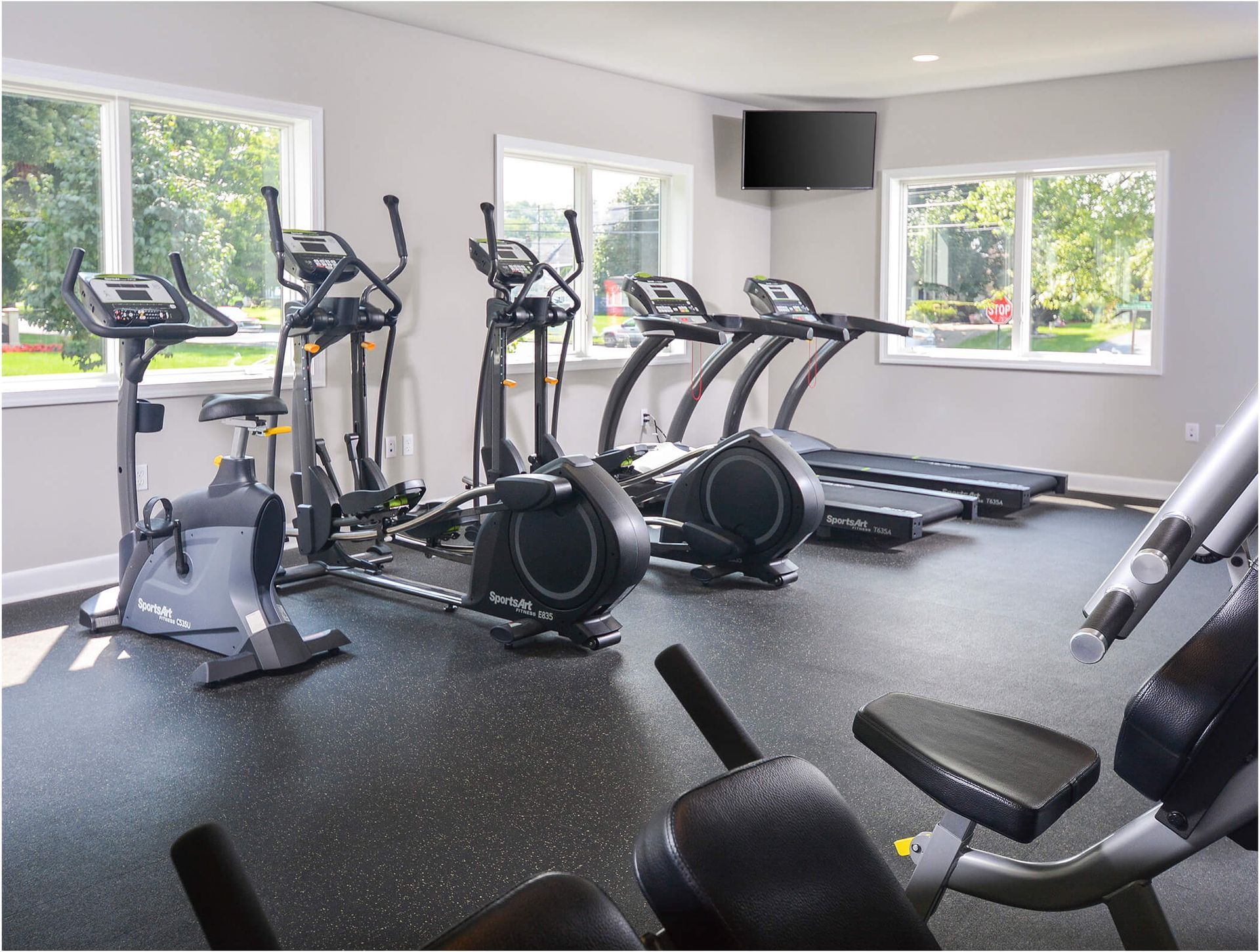 Bright apartment gym with treadmills, ellipticals, and stationary bikes beside large windows.
