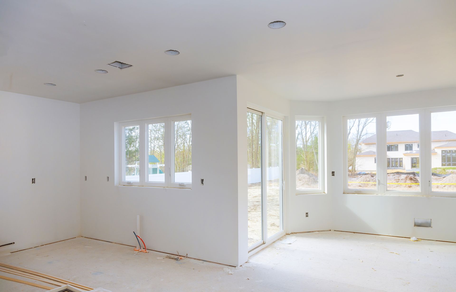Bright unfinished room with white walls, recessed lights, and large windows overlooking a neighborhood