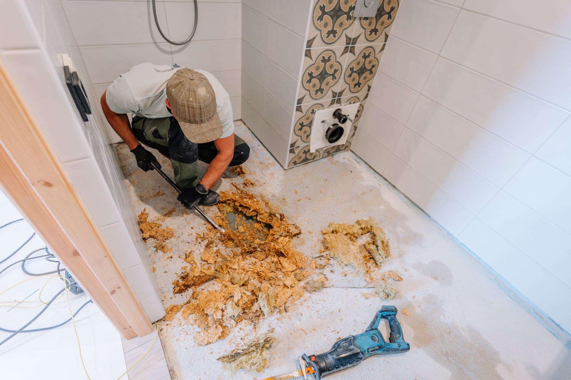 Person cleaning debris from a bathroom floor with a shovel and bucket, white tiled walls and floor.
