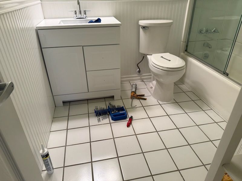Small white-tiled bathroom with toilet, sink cabinet, and scattered cleaning tools on the floor.