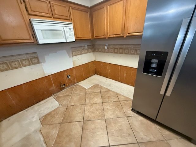 Empty kitchen corner with tan tile floor, wood cabinets, white microwave, and stainless steel refrigerator.