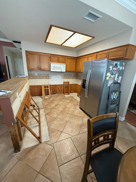 Bright kitchen with wood cabinets, stainless fridge, tiled floor, and a small breakfast counter.