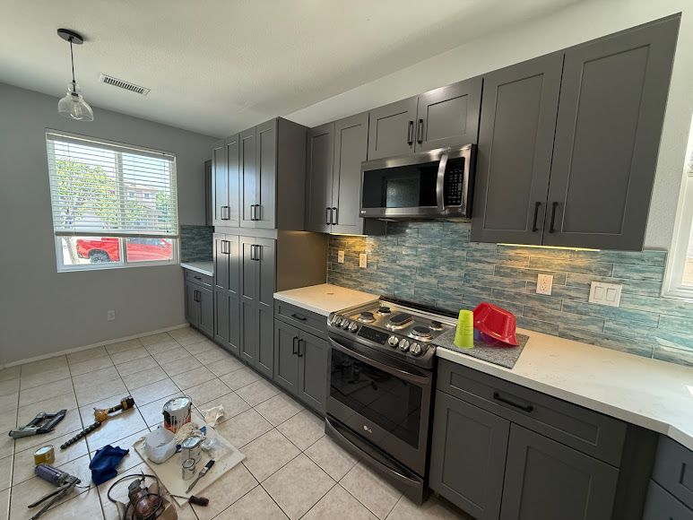 Modern gray kitchen with tiled floor, stainless appliances, and cleaning supplies on the floor near the window.