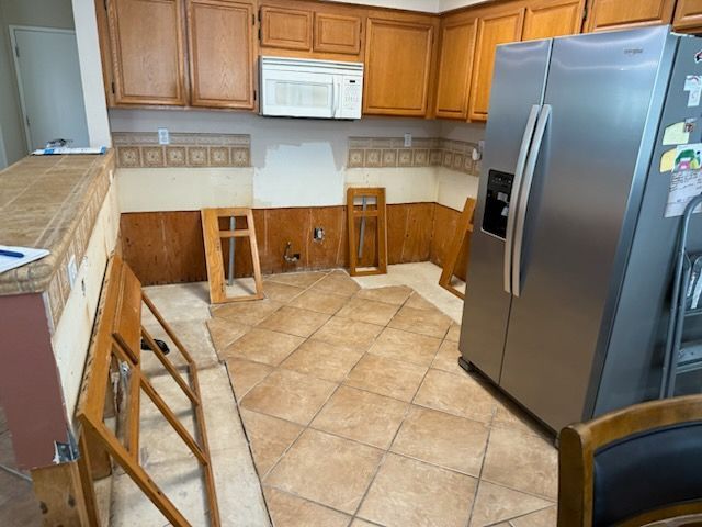 Kitchen with tan tile floor, wooden cabinets, white microwave, and stainless steel refrigerator; chairs are stacked nearby