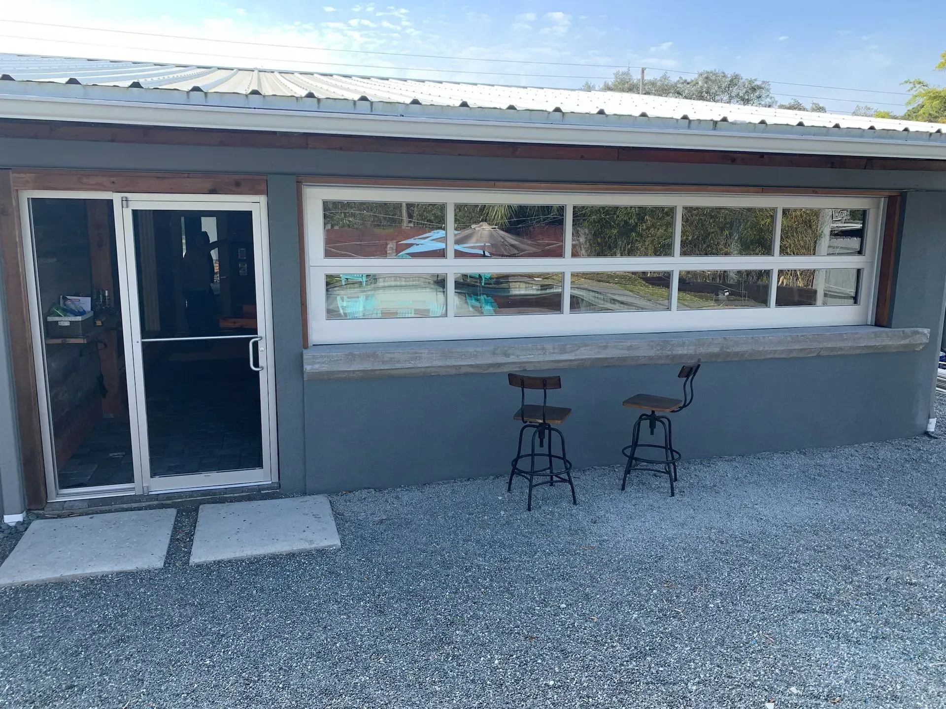 Gray building with door and large window. Two stools sit outside on a gravel patio.