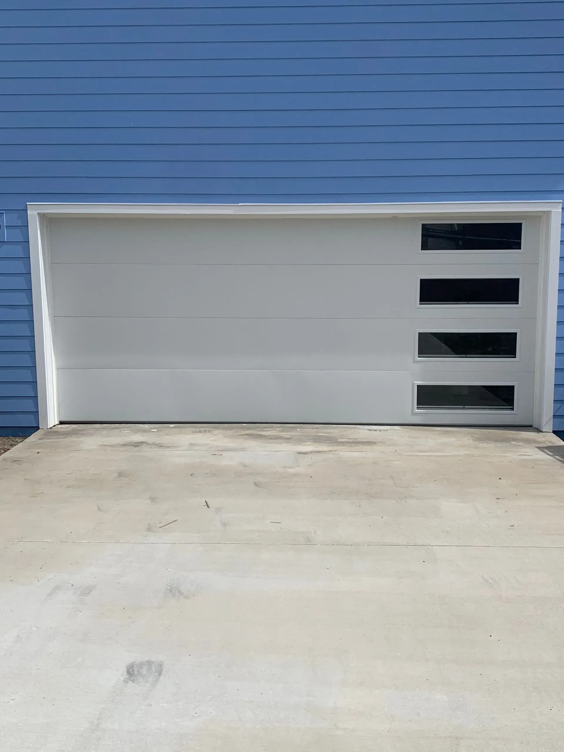 White garage door with four rectangular windows, on blue siding. Concrete driveway.