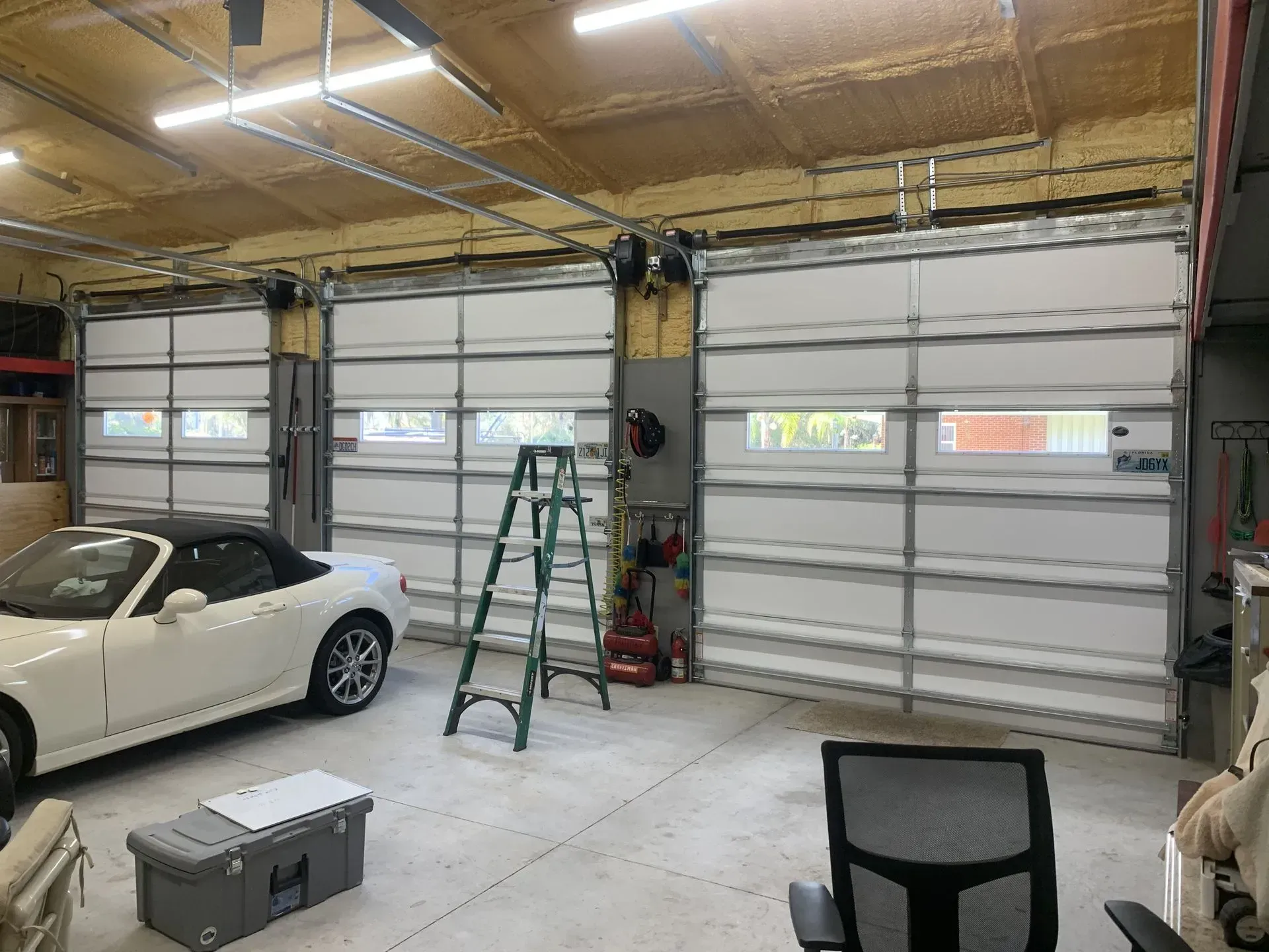 Three closed white garage doors, one white car inside a garage. A ladder and chair are also in the garage.